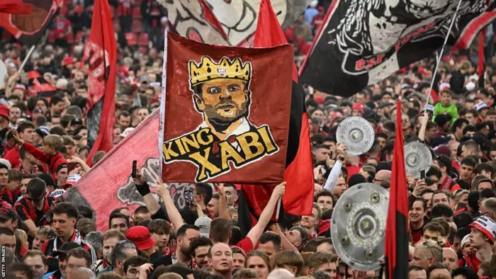 Pendukung Leverkusen yang membentangkan bendera bergambar Xabi Alonso sebagai tanda penghormatan (Sumber Foto : Getty Images)