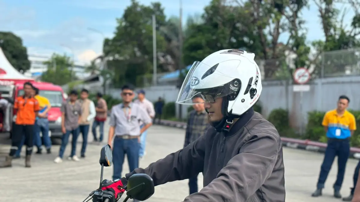 Seorang pengendara motor mengenakan helm dan sarung tangan saat bersiap berkendara, disaksikan sejumlah orang di area terbuka.