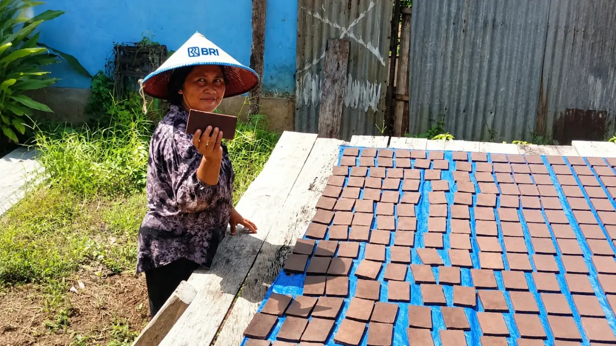 Seorang pelaku usaha mikro menunjukkan buku tabungan BRI di tengah aktivitas produksi usaha rumahan, dengan hasil olahan yang sedang dijemur di area terbuka.