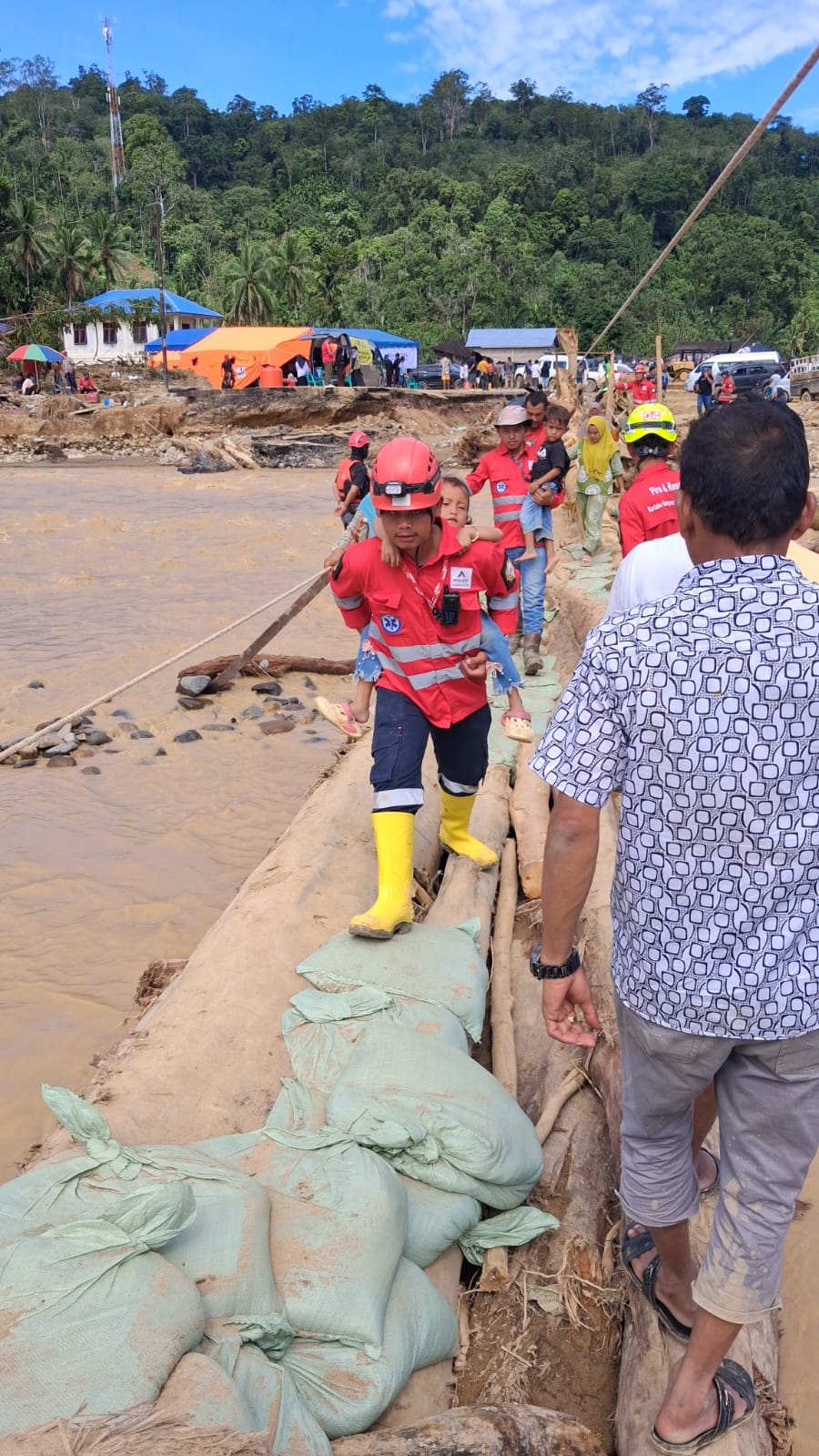 Anggota Tim Tanggap Darurat (ERT) Agincourt Resources menggendong seorang anak meniti jembatan darurat yang menghubungkan Tapanuli Selatan dan Tapanuli Tengah, Selasa (2/12/2025).