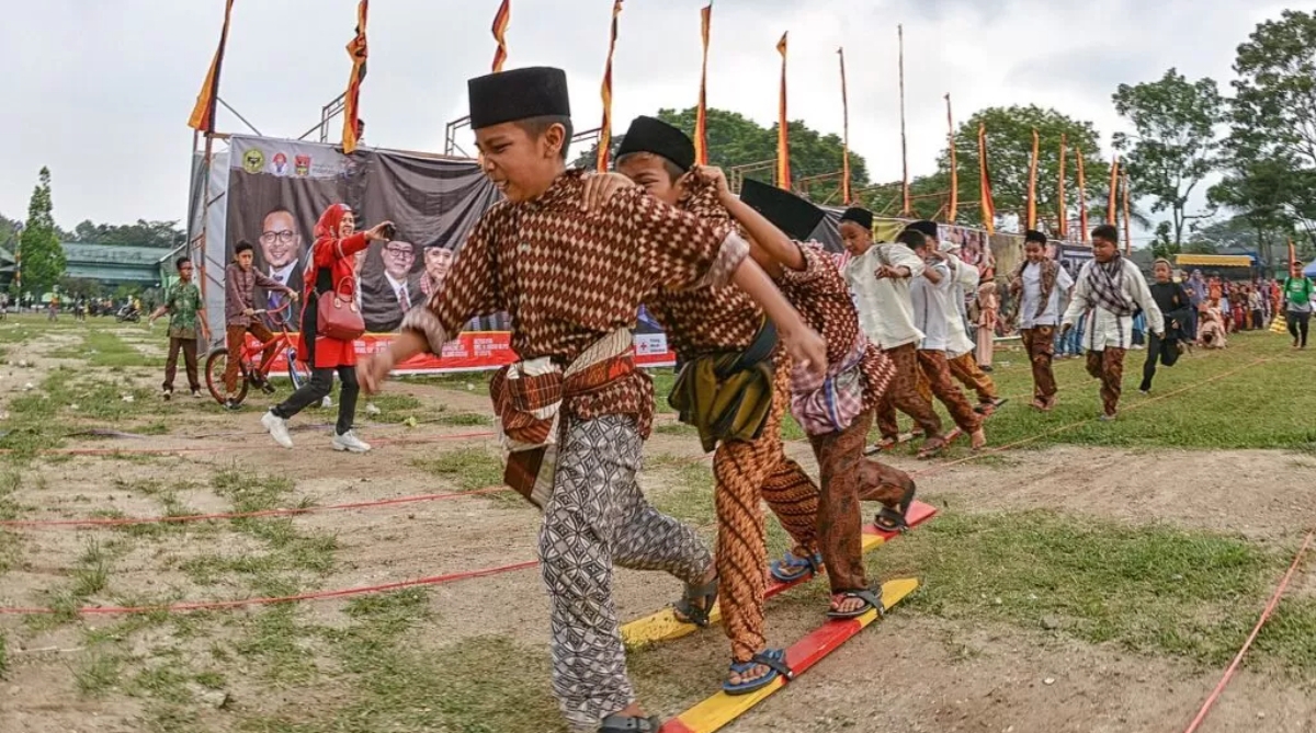 Anak-anak mengenakan busana tradisional Jawa mengikuti lomba bakiak dalam rangkaian perayaan HUT ke-79 Kemerdekaan RI di sebuah lapangan desa, penuh semangat dan sorak sorai penonton. (Foto: Jawa Pos)