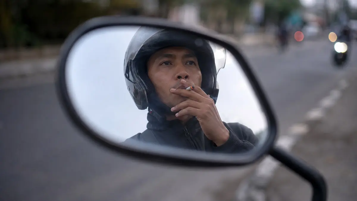 Ilustrasi. Pengendara sepeda motor merokok saat berkendara di jalan raya. Kebiasaan tersebut dapat mengganggu konsentrasi dan membahayakan pengguna jalan lain. (ChatGPT)