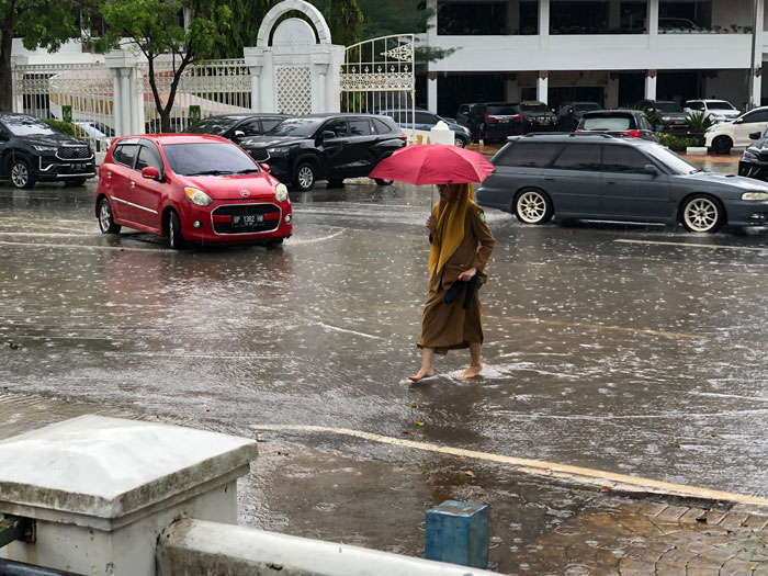 Seorang pegawai Pemko Batam terpaksa melepas sepatu demi melintas jalan. Hujan, jalan tergenang.