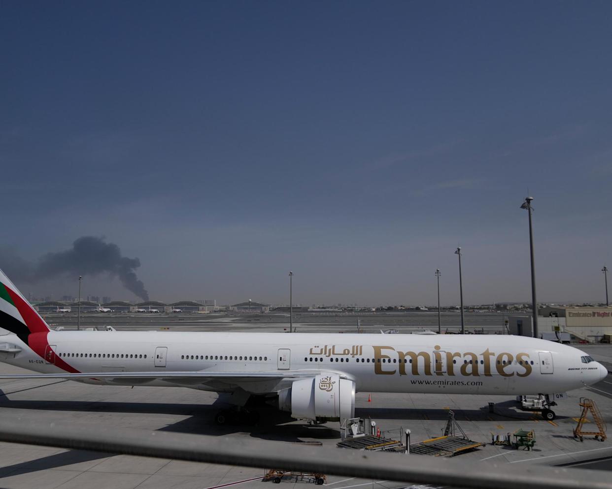 Kepulan asap akibat serangan Iran terlihat di belakang sebuah pesawat Emirates yang terparkir di Bandara Dubai, Minggu (1/3/2026). Ribuan penerbangan telah dibatalkan sejak AS serang Iran.