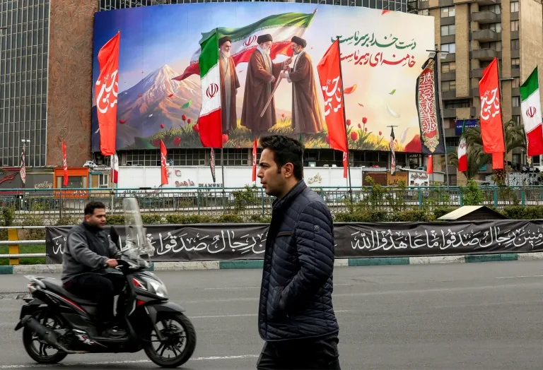 Warga Iran berjalan di depan baliho Mojtaba Khamenei di Teheran. Baliho itu simbol penyerahan bendera nasional dari ayahnya yang tewas terbunuh oleh tentara AS dan Israel pada awal perang. F AFP