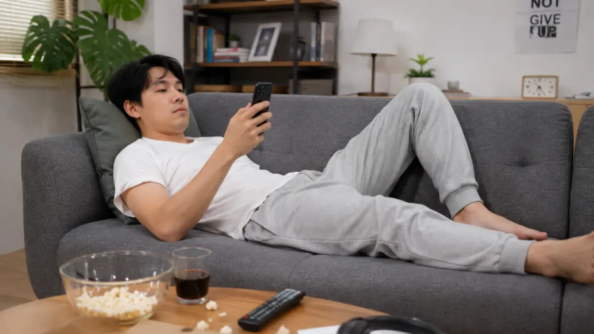 Ilustrasi seorang pria yang sedang bersantai di sofa sambil menggunakan ponsel.