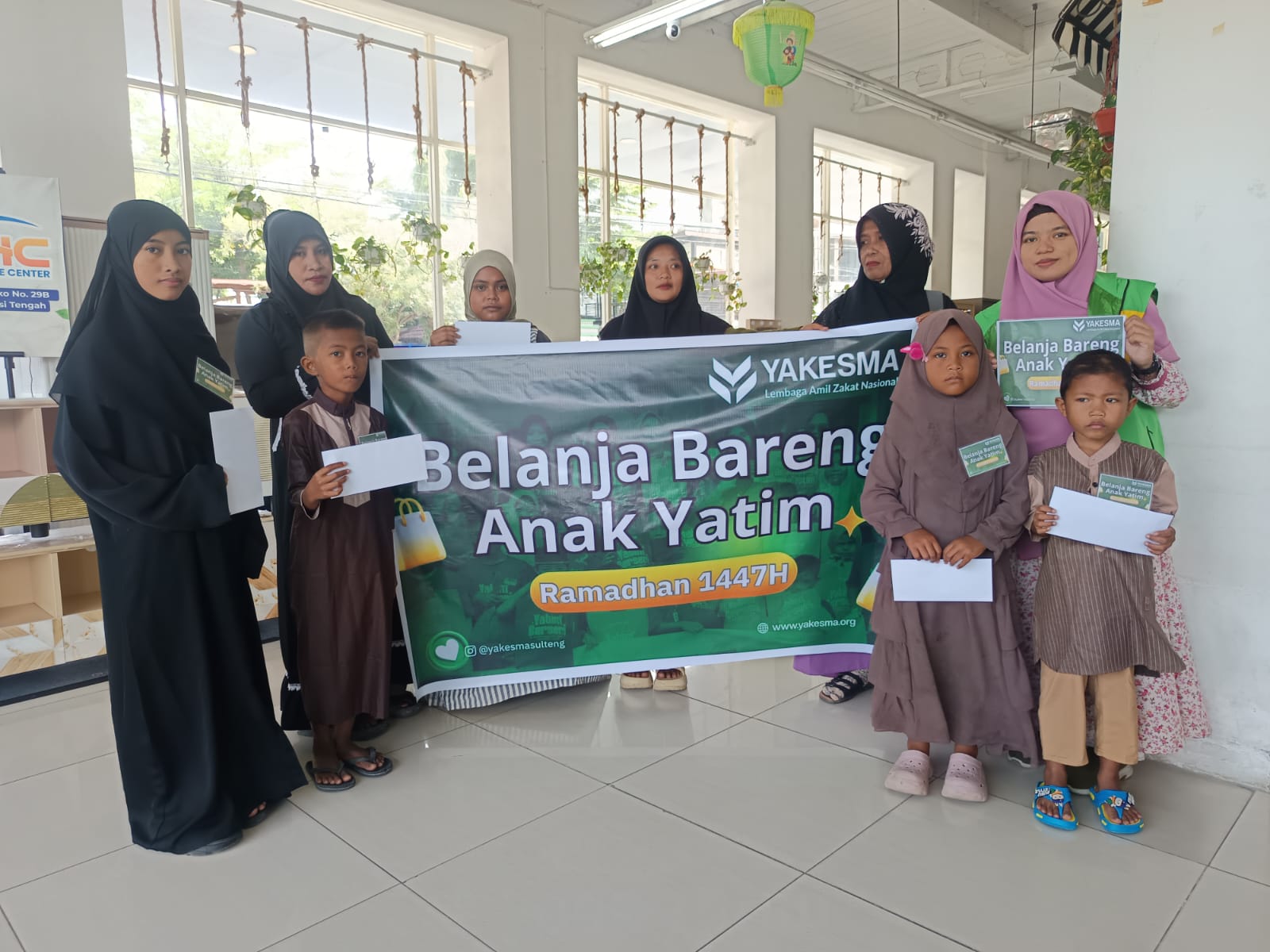 Anak-anak yatim berpose bahagia bersama pendamping dan relawan YAKESMA Sulawesi Tengah, memegang plakat dan banner "Belanja Bareng Anak Yatim Ramadan 1447 H".