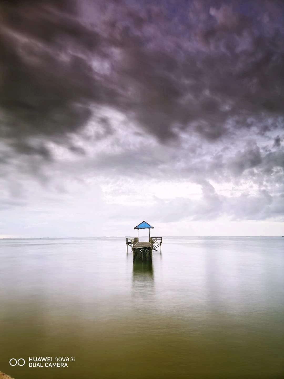 Pengunjung duduk santai di tepi Pantai AURI Balikpapan saat senja, dengan latar langit jingga dan pepohonan rindang di sekitar.