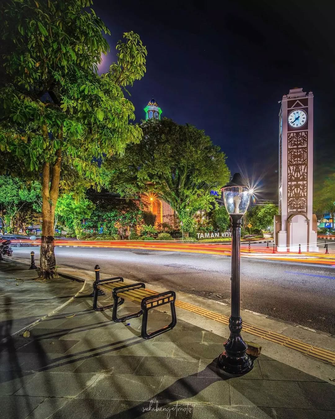 Foto Tugu Jam Taman Kota Balikpapan di malam hari dengan cahaya lampu warna-warni yang memantul di taman hijau &mdash; simbol waktu dan keindahan kota.