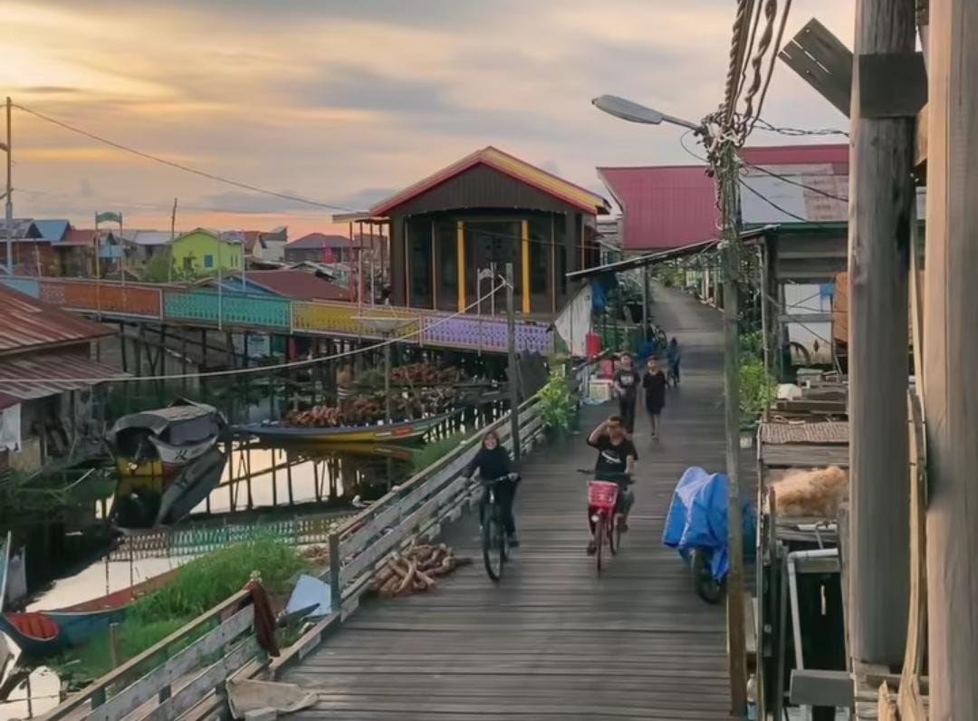 Pemandangan sore di perkampungan nelayan Muara Enggalam dengan rumah panggung warna-warni dan anak-anak bersepeda.
