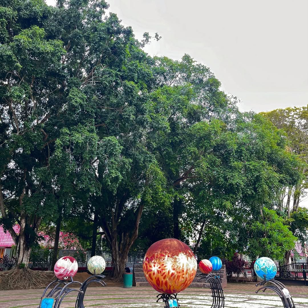 Deretan replika planet berwarna cerah berdiri di area terbuka Planetarium Jagad Raya Tenggarong, dikelilingi pepohonan rindang yang menambah suasana sejuk dan edukatif.