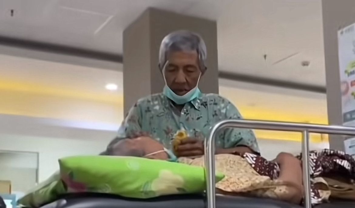 Kakek menyuapi istrinya yang sakit dengan tatapan penuh kasih &mdash; visual hangat yang menggambarkan cinta sejati di usia senja.