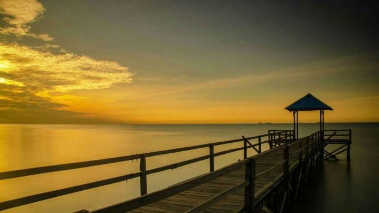 Pantai Sepinggan Balikpapan &mdash; siluet dermaga kayu dengan latar langit jingga yang memantul di permukaan laut tenang.