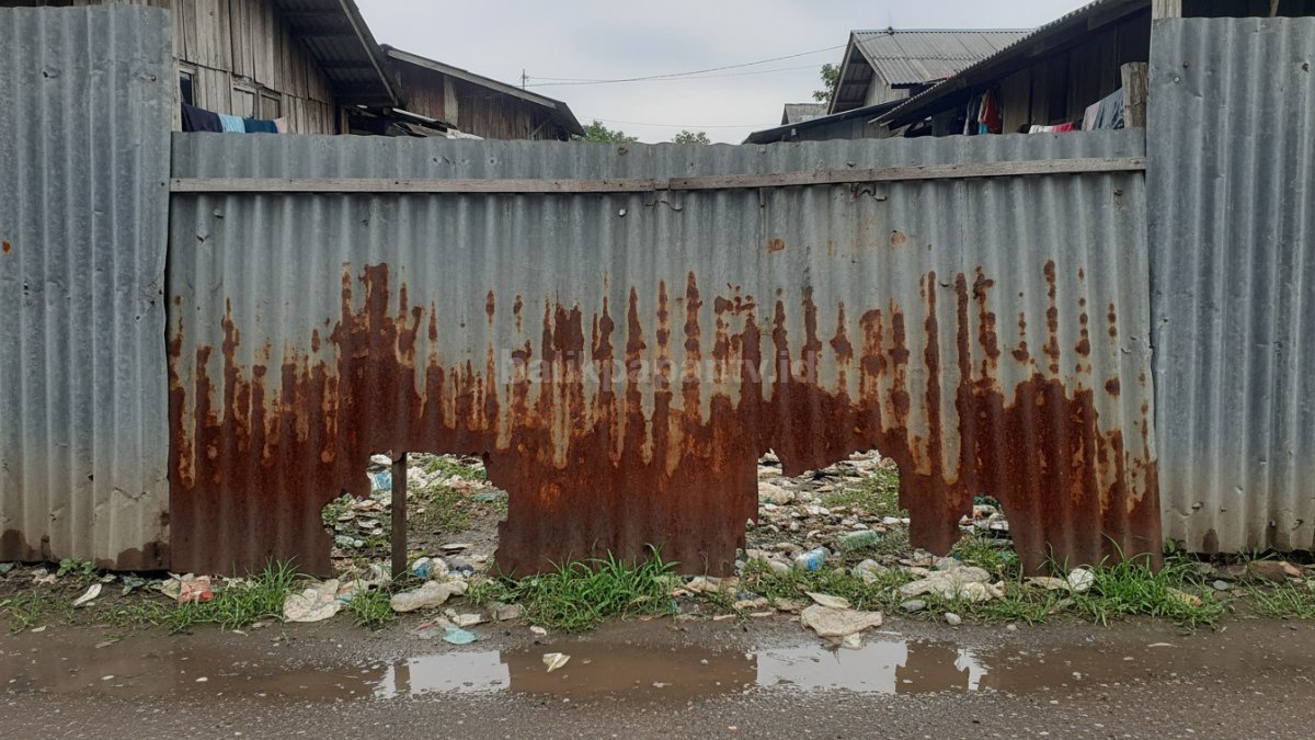 Seng berkarat dan berlubang di bagian bawah pagar, memperlihatkan dampak tanpa perawatan