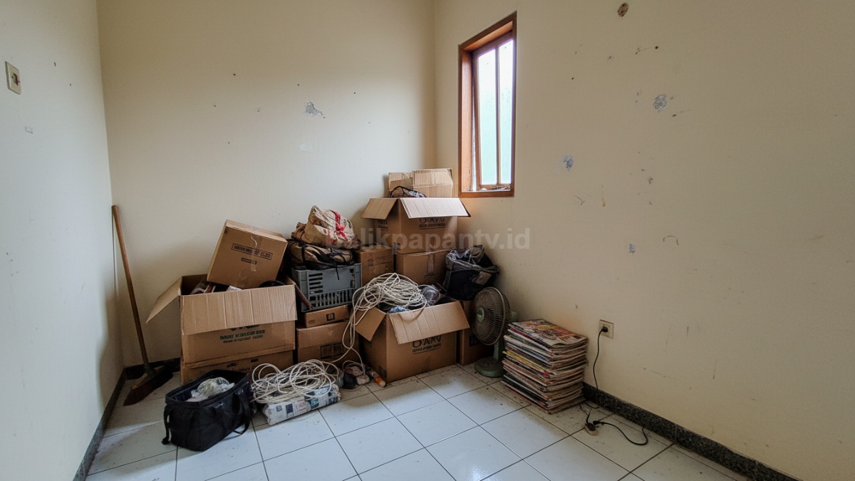Pojok ruangan di rumah kontrakan berisi tumpukan kardus dan barang lama tanpa penataan jelas