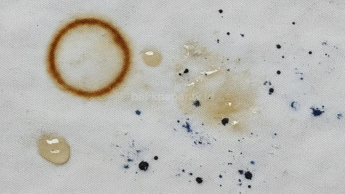 Close-up kain putih dengan berbagai jenis noda seperti kopi, minyak, dan tinta yang terlihat jelas menempel di serat kain.