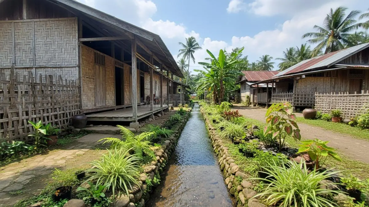 Tampilan sebuah rumah sederhana di Indonesia dengan area pinggir selokan yang ditata rapi menggunakan berbagai tanaman hias tahan air seperti lili paris, pakis, dan caladium. Selokan terlihat bersih dengan aliran air lancar, di sampingnya terdapat susunan batu kecil dan tanaman hijau yang tumbuh subur, menciptakan suasana adem, alami, dan enak dipandang.