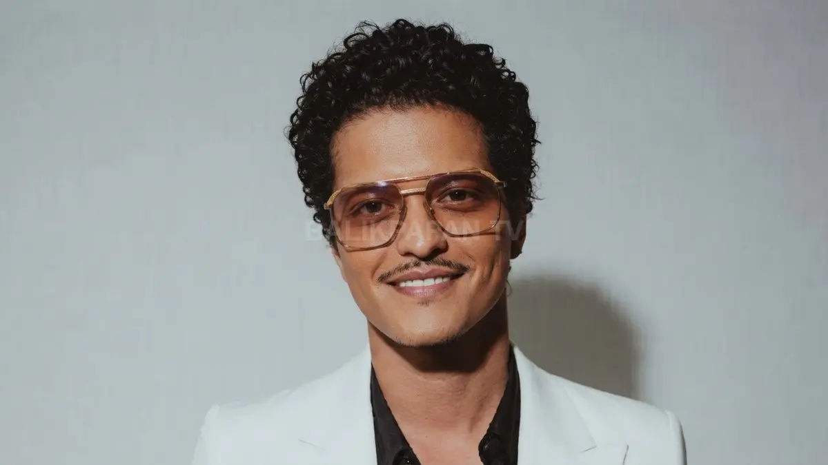 Potret kiki Bruno Mars berambut keriting dengan kacamata berbingkai emas dan setelan jas putih. (BTV/Ai)