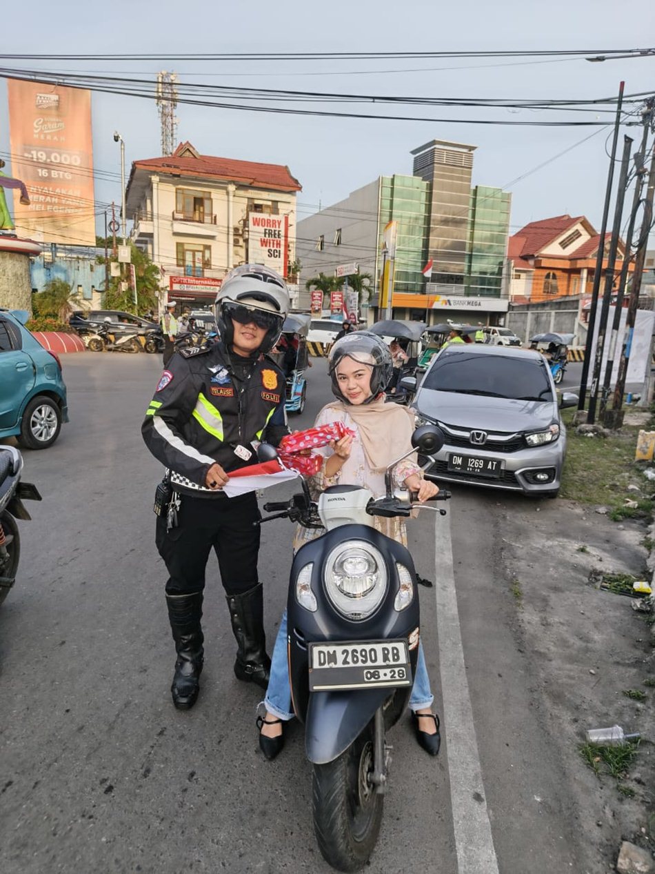 Personel Satlantas Polresta Gorontalo Kota berdiri dengan membawa bendera merah putih mini dan bingkisan hadiah kepada pengendara yang berhenti di lampu merah.(F:hms-Pol)