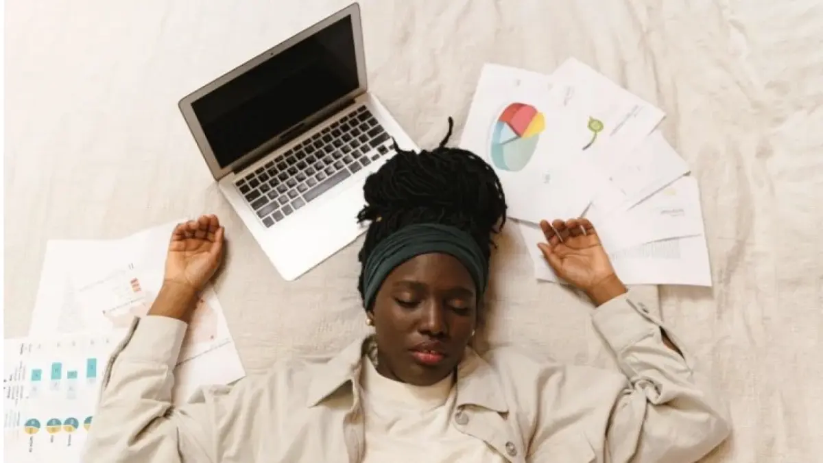 Seorang wanita yang kelelahan memejamkan mata sembari berbaring di samping laptop dan tumpukan kertas setelah bekerja. (Pexels-Antoni Shkraba Production)