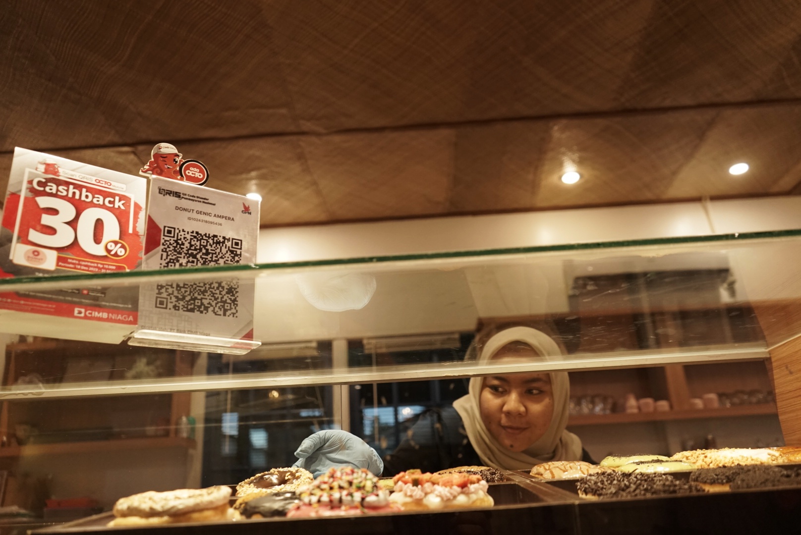 PROMO: Pelayan Kafe Donutsgenic di Jl Ampera, Pontianak tengah menyusun donat di etalase. Sebuah papan promo transaksi nontunai QRIS dari Octo Pay CIMB Niaga terpampang di atasnya.