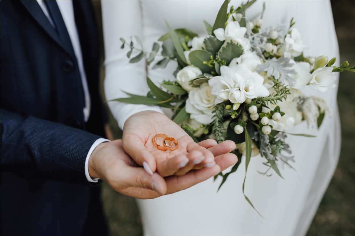 Ilustrasi sepasang pengantin memegang cincin pernikahan dengan latar bunga sederhana, melambangkan pernikahan yang berkesan meski dilakukan secara hemat dan penuh makna.