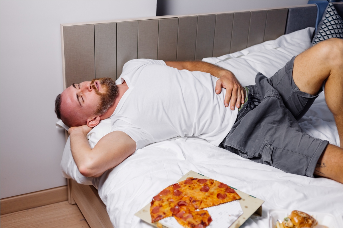 Ilustrasi seorang pria berbaring di tempat tidur setelah makan dengan piring makanan di sampingnya, menggambarkan kebiasaan tidur setelah makan yang dapat berdampak pada kesehatan pencernaan.