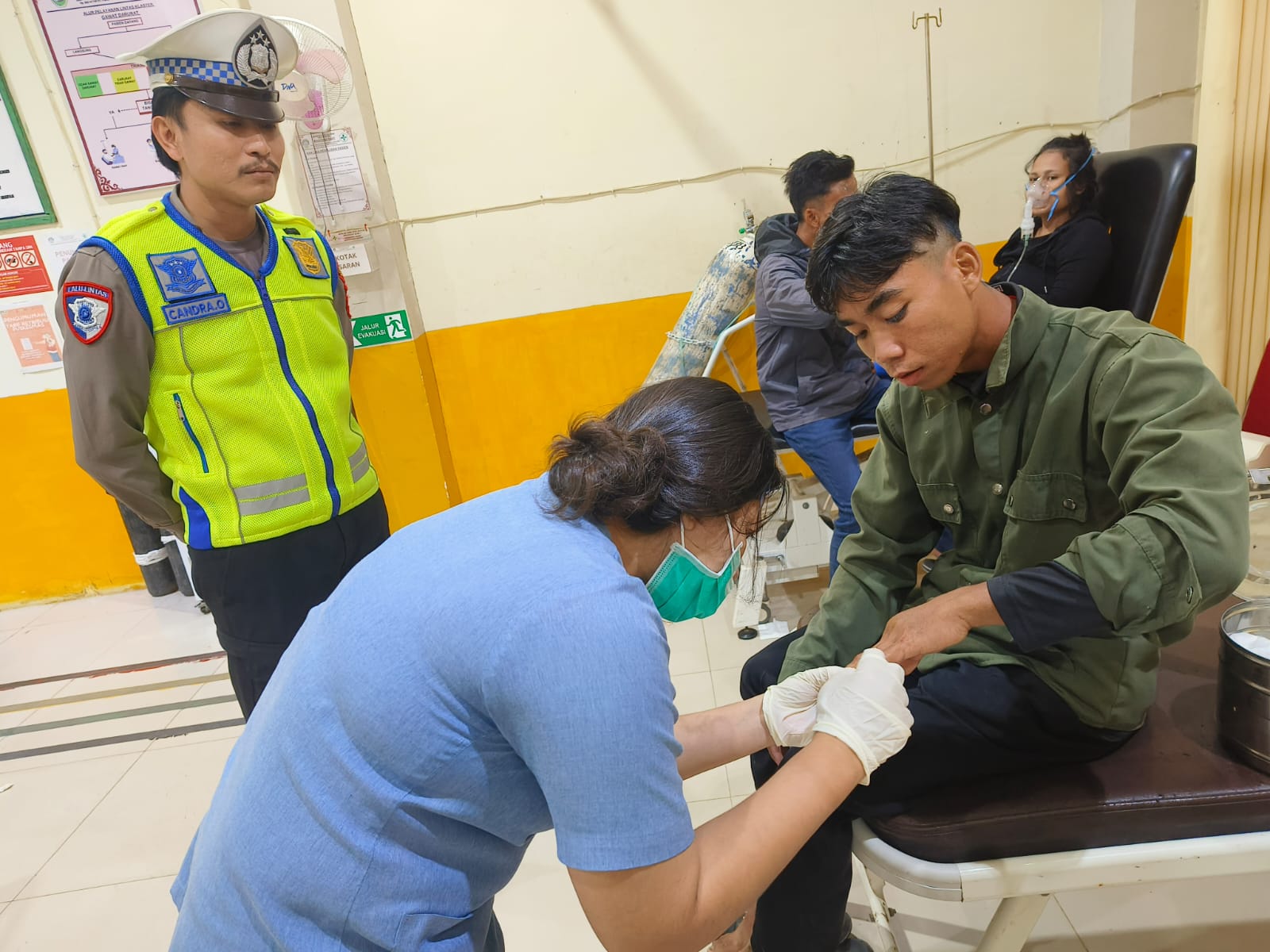 Petugas medis memberikan perawatan kepada korban kecelakaan lalu lintas di fasilitas kesehatan, disaksikan anggota kepolisian yang melakukan pendataan kejadian.