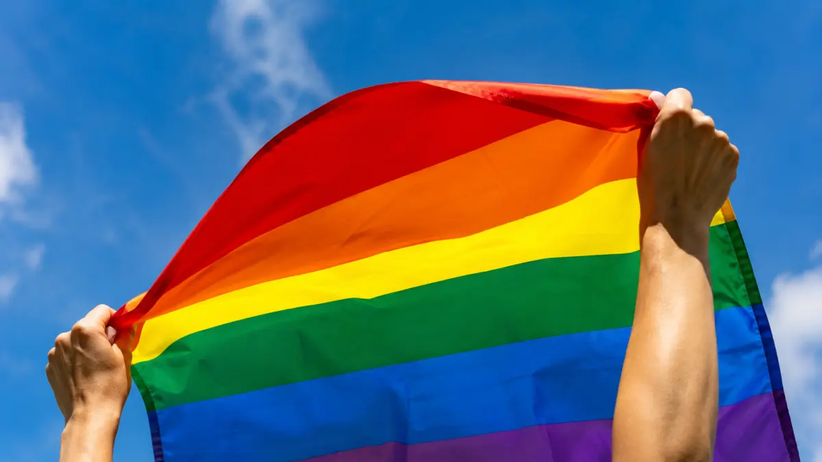 Tangan seseorang merentangkan bendera pelangi di ruang terbuka dengan latar langit biru cerah sebagai simbol LGBTQ.  Presiden Prabowo Subianto menerbitkan Perpres Nomor 111 Tahun 2025 yang mencantumkan penyebaran perilaku LGBTQ sebagai salah satu ancaman nonmiliter terhadap pertahanan negara.