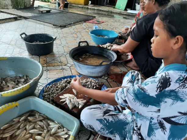 Ibu-ibu hingga remaja putri sedang membelah ikan bilis segar yang akan diolah menjadi ikan asin. Ikan Asin Kak Ros menjadi UMKM yang menyokong ekonomi warga sekitar.