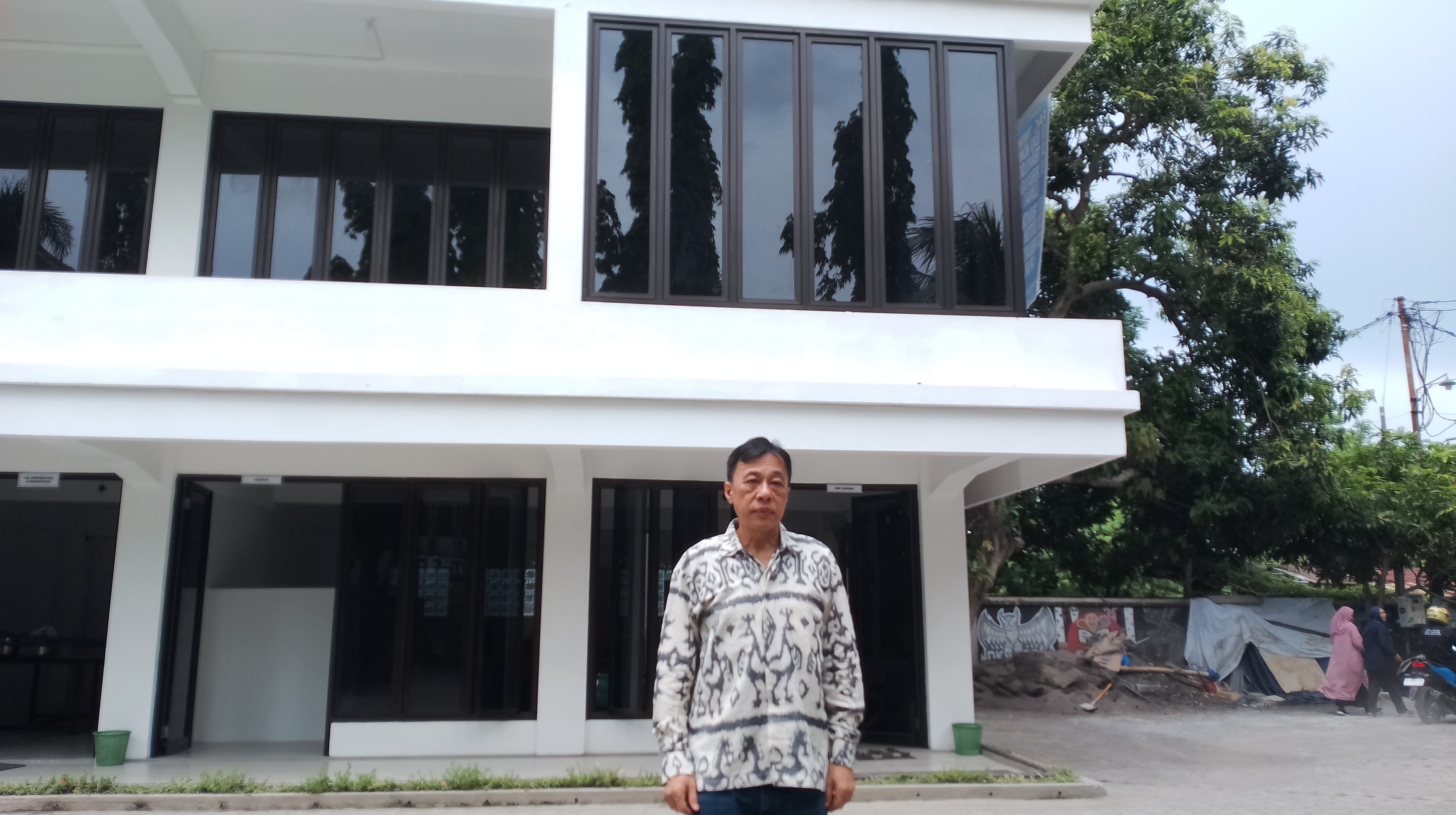 Rektor Universitas Bumigora Dr.Ir. Anthony Anggrawan, Ph.D berdiri dengan latar belakang foto gedung baru Fakultas Kedokteran.