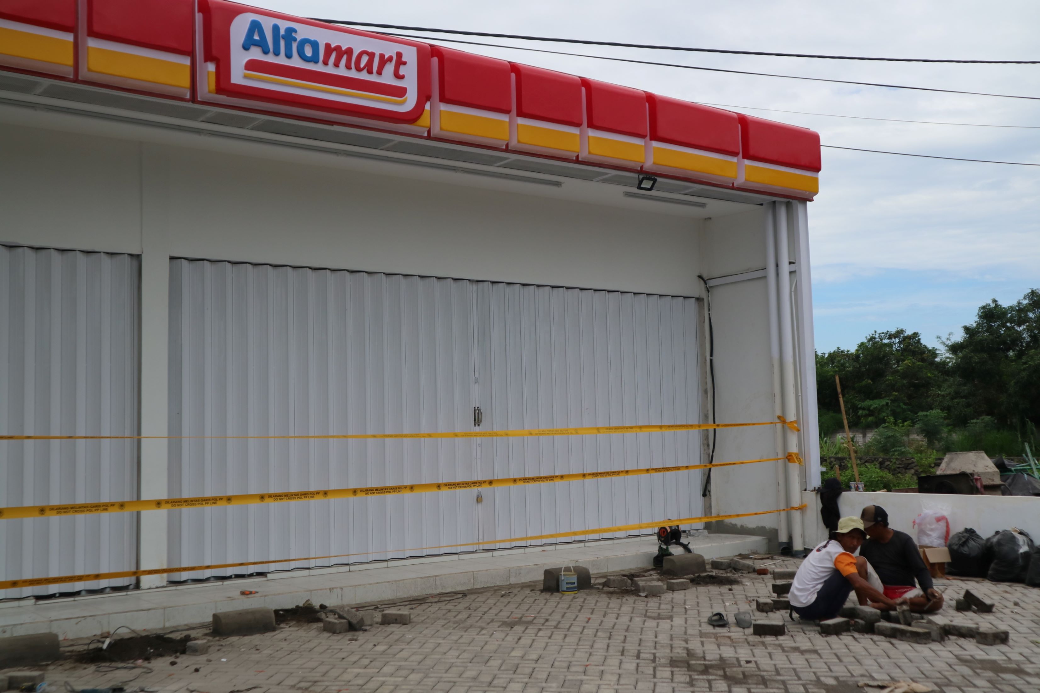 TAK BERIZIN: Sejumlah buruh tukang sedang memasang paving block di depan area toko Alfamart yang terpasang pita kuning larangan melintas oleh Satpol PP Loteng di Loteng.