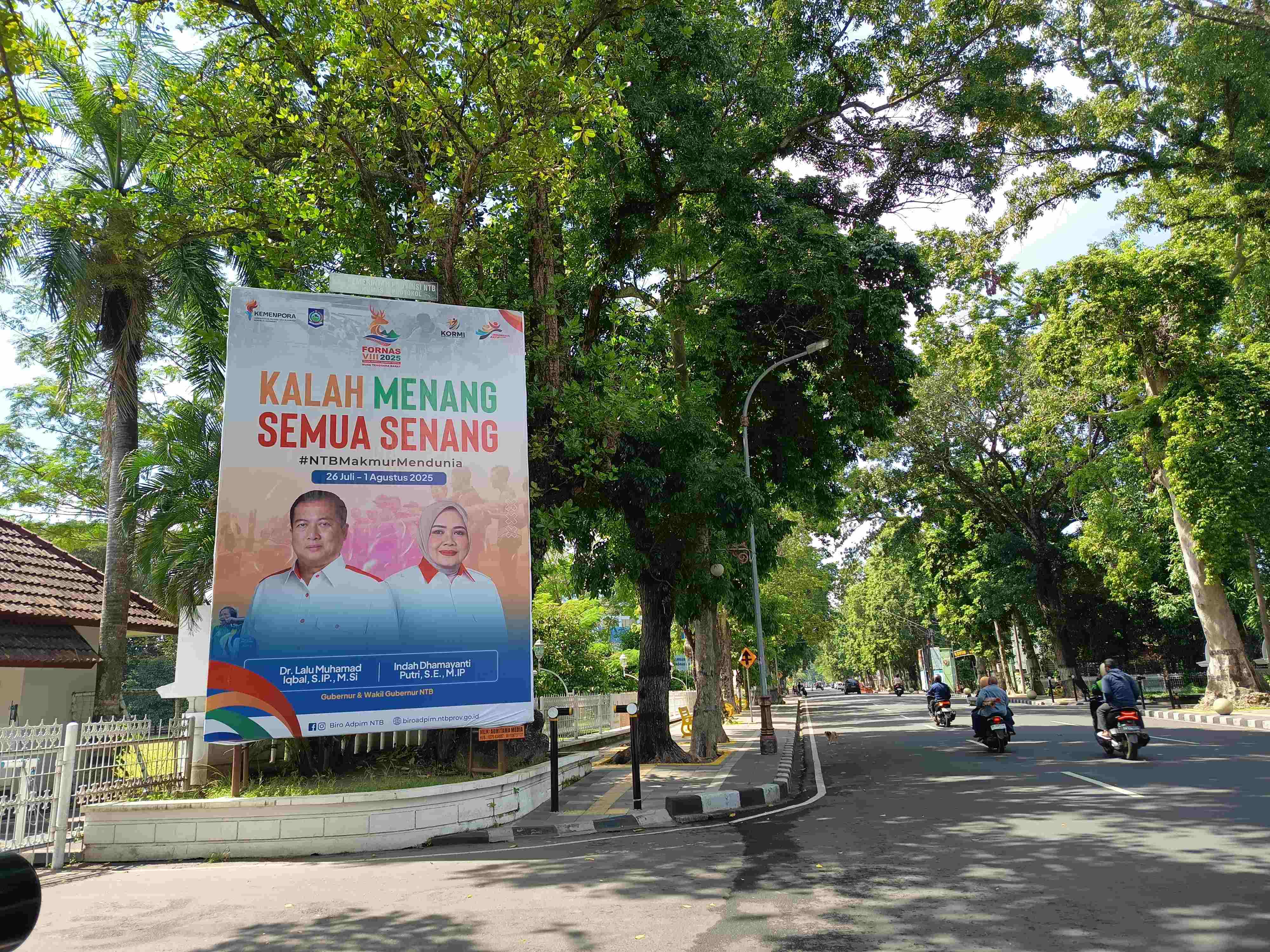 GAWE NASIONAL: Pengendara sepeda motor melintas baliho FORNAS VIII NTB 2025 terpampang di depan Kantor Gubernur NTB.