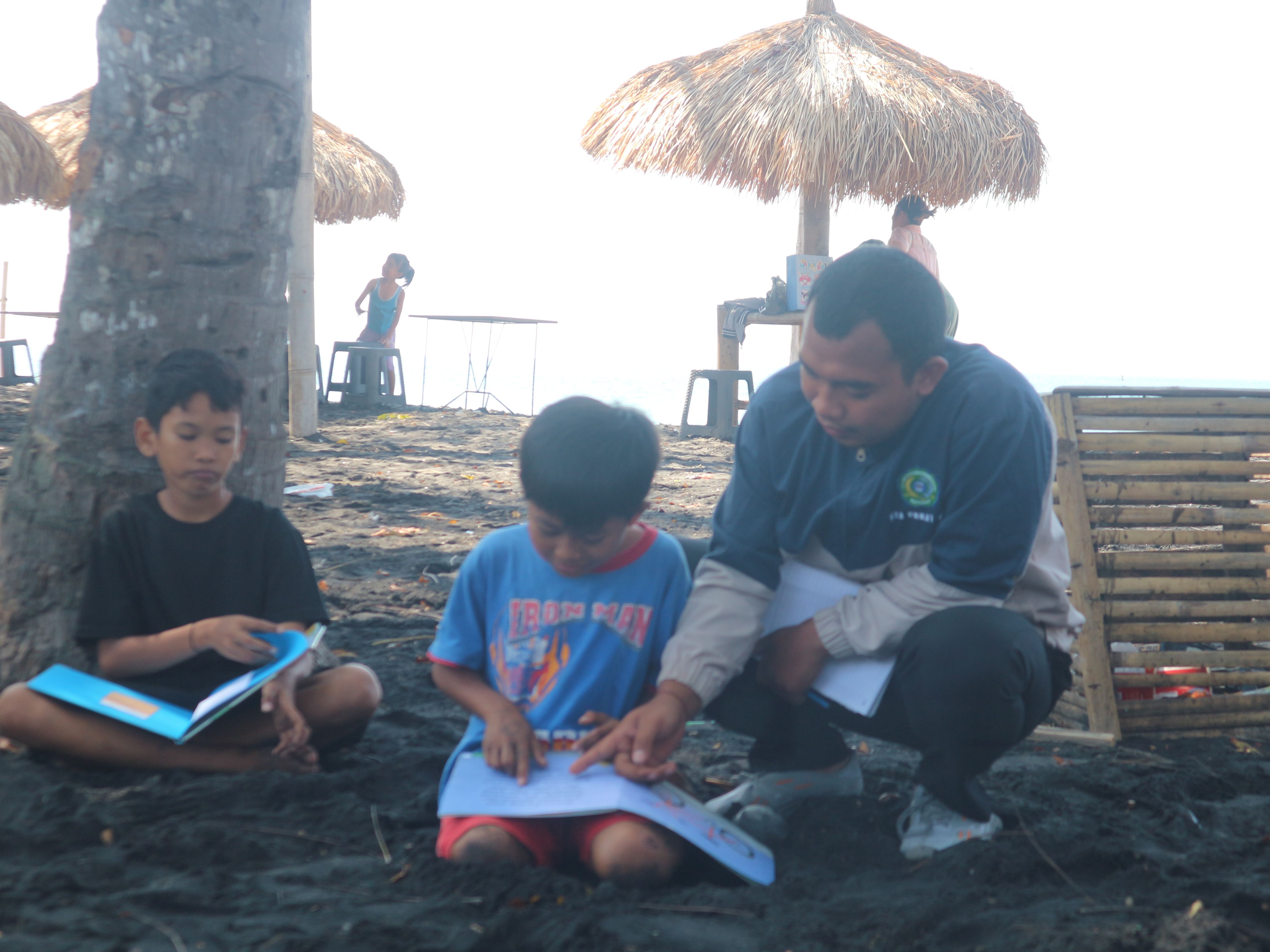 Salah seorang pemuda sedang membimbing anak-anak dusun perenang membaca buku di Pantai Sunrise Land Lombok