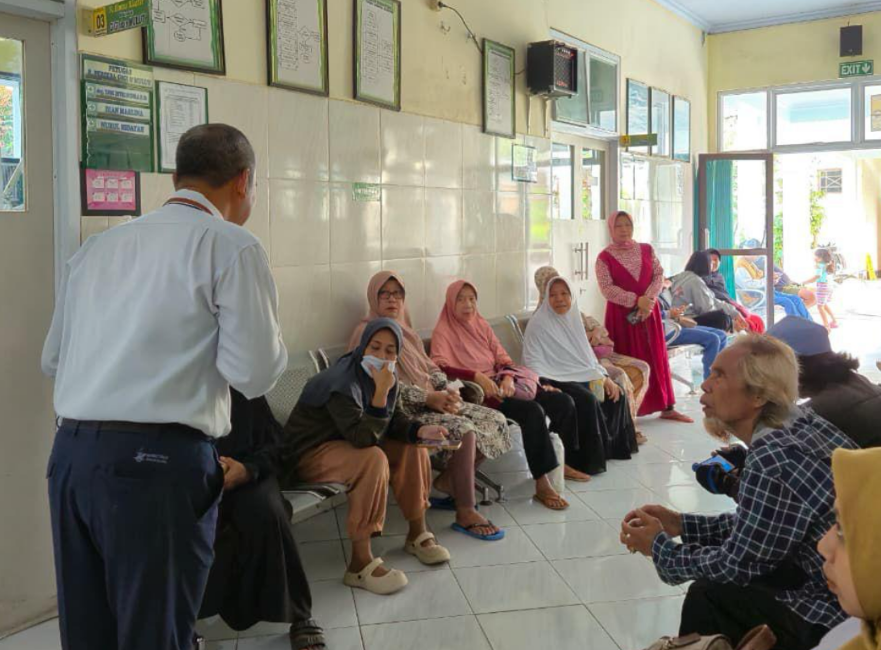 Manager Hari Ini (kemeja putih berdasi) memegang Manager Hari Ini dengan kemeja putih berdasi biru tersenyum sambil memantau dan menyapa pasien di ruang tunggu puskesmas Kota Mataram.