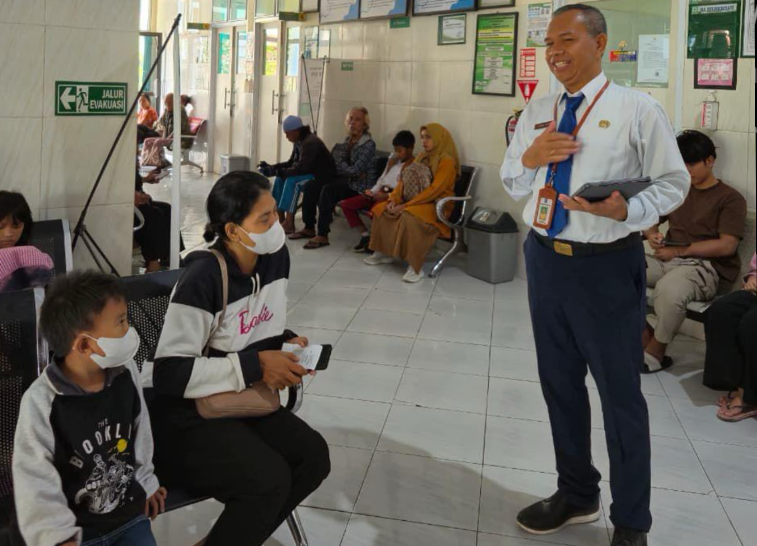 Manager Hari Ini dengan kemeja putih berdasi biru tersenyum sambil memantau dan menyapa pasien di ruang tunggu puskesmas Kota Mataram.