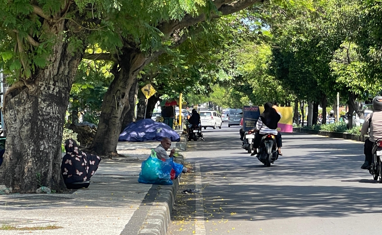 Seorang warga duduk di pinggir jalan sambil sesekali melambaikan tangan meminta belas kasih dari pengendara yang melintas. 
