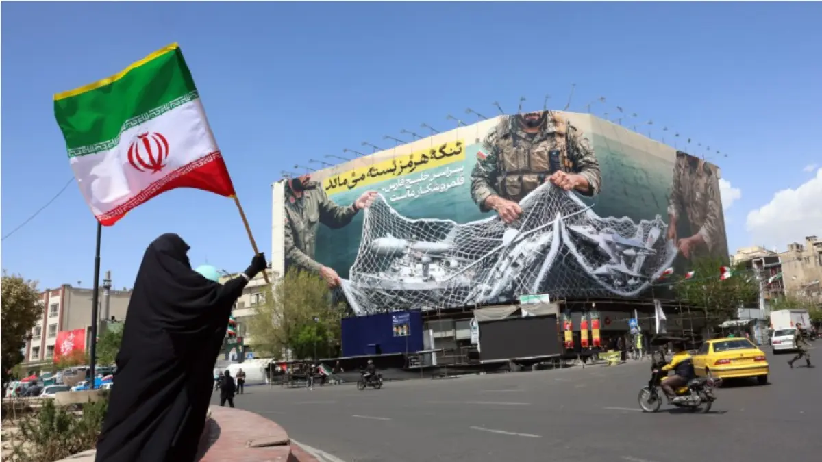 SELAT HORMUZ: Seorang wanita memegang bendera Iran sambil berdiri di dekat papan reklame dengan kalimat bertuliskan ‘Selat Hormuz tetap ditutup’ di Lapangan Enqelab di Teheran, Ahad (5/4). (AFP)