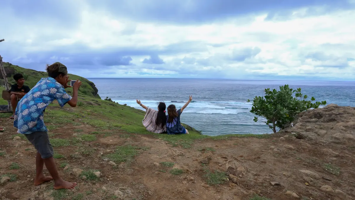 Seorang anak sedang memotret dua pengunjung yang berswafoto pemandangan Pantai Tanjung Aan dari atas bukit Merese di Lombok Tengah, belum lama ini. (Ivan/Lombok Post) 
