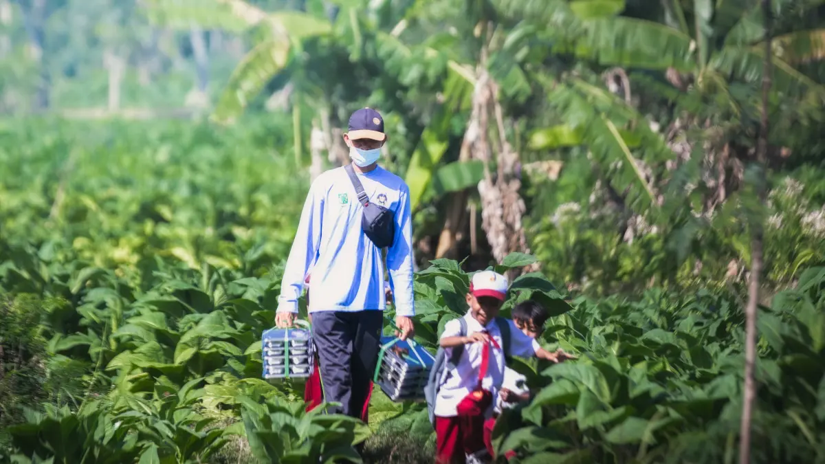 SALURKAN MBG: Seorang petugas SPPG membawa ompreng berisi MBG, dengan berjalan kaki menjangkau sekolah di pelosok Lombok Tengah (Loteng), beberapa waktu lalu. (IVAN/LOMBOK POST).