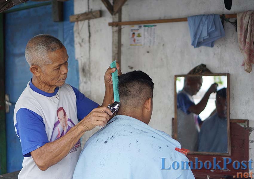 TUKANG CUKUR: Muhammad, tukang cukur konvensional dari Rempung sedang memangkas rambut pelanggannya di perempatan Desa Rempung, Kecamatan Sukamulia, belum lama ini.