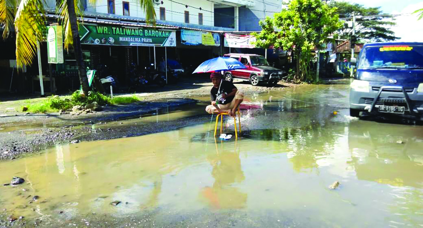 PROTES: Amaq Ohan, warga Tengari, Praya, Lombok Tengah, sedang memancing ikan di kubangan air di jalan berlubang di perempatan kampus IPDN, Selasa (16/3/2021). (Dedi/Lombok Post)