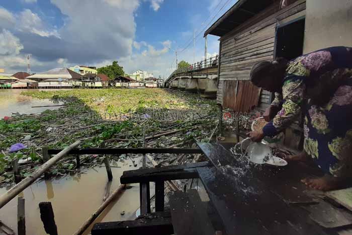 TERCEMAR: Warga di bantaran Sungai Martapura mencuci piring. Tampak di belakang, tumpukan sampah dan gulma menyumbat kolong Jembatan 9 November. | Foto: Wahyu Ramadhan/Radar Banjarmasin
