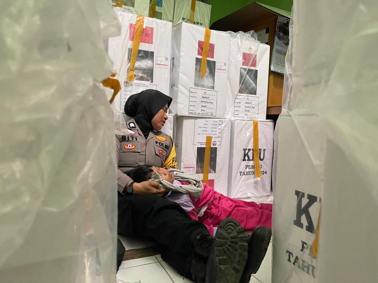 Personel Polsek Perhentian Raja Brigadir Siti Fatimah Yulis berjibaku menjaga logistik pasca pendistribusian dikunjungi anaknya yang masih belia di aula Kantor Camat Perhentian Raja, Senin (12/2/2024)
