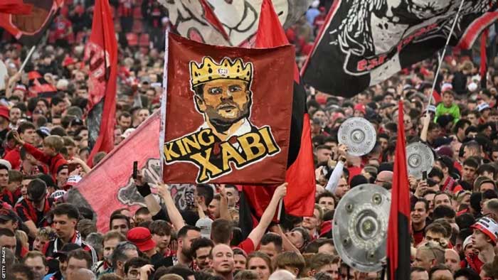 Pendukung Leverkusen yang membentangkan bendera bergambar Xabi Alonso sebagai tanda penghormatan