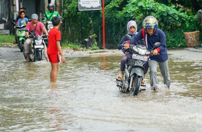 Seorang pengendara mendorong sepeda motor yang mogok saat melewati genangan banjir di Jalan Cipta Karya, Ahad (9/6/2024).