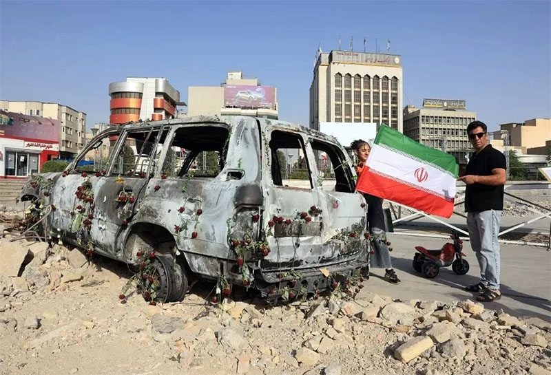 Seorang pria mengibarkan bendera Iran di samping ambulans Bulan Sabit Merah Iran yang hancur akibat serangan udara Israel.