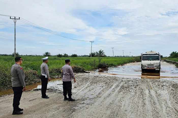 Personel Polsek Bonai Darussalam memantau kondisi ruas jalan provinsi Sontang batas Duri tergenang air saat dilintasi truk pengangkut sawit, Kamis (19/2/2026).